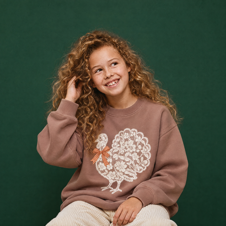 A young girl with long curly light brown hair smiles while wearing a brown crewneck sweatshirt with a white lace turkey graphic and an orange bow, set against a solid dark green background.